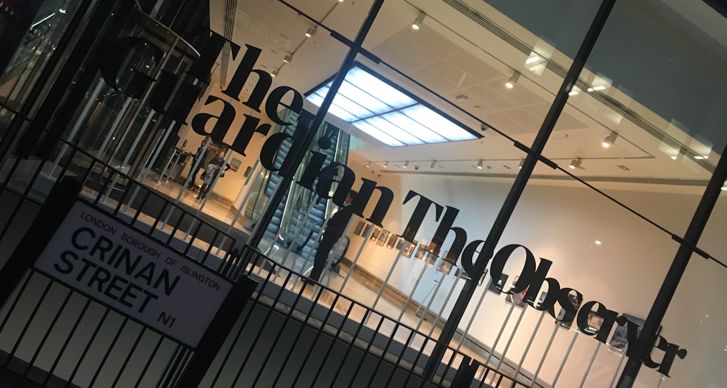 A modern glass building entrance with large black lettering spelling out a newspaper name on the glass. The interior appears well-lit with a visible staircase, minimalistic design, and a sign with a street name in the foreground.