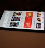 A smartphone displaying a streaming app interface, showing various movie and TV show thumbnails. The screen includes categories like 'For you' and 'New release hit films', with images from popular films. The background is a dimly lit setting with a fabric surface.