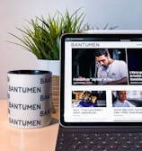 A tablet displaying a magazine website is positioned on a table, next to a mug with the word 'Bantumen' printed multiple times and a small potted plant. The tablet shows various articles and images on the screen.