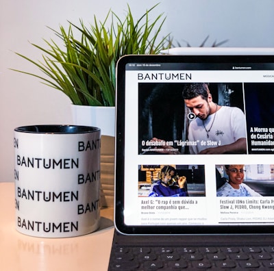 A tablet displaying a magazine website is positioned on a table, next to a mug with the word 'Bantumen' printed multiple times and a small potted plant. The tablet shows various articles and images on the screen.