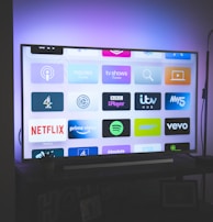 A flat-screen television is displayed in a dimly lit room, showcasing a variety of streaming service apps on its screen, such as Netflix, Prime Video, and Spotify. The TV is positioned on a black stand beside a tall floor lamp, casting a soft ambient light on the surrounding area.