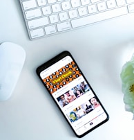 A smartphone displaying a social media or entertainment app, placed on a white desk. Nearby, there is an Apple Magic Keyboard and a white mouse. A partially visible white flower is in the top right corner.