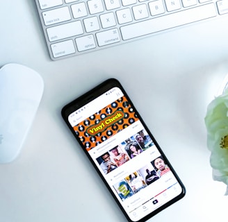A smartphone displaying a social media or entertainment app, placed on a white desk. Nearby, there is an Apple Magic Keyboard and a white mouse. A partially visible white flower is in the top right corner.