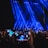 A large crowd of people holding up smartphones with their screens lit, capturing or recording an entertainment event on a stage illuminated by prominent blue spotlights.