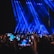 A large crowd of people holding up smartphones with their screens lit, capturing or recording an entertainment event on a stage illuminated by prominent blue spotlights.