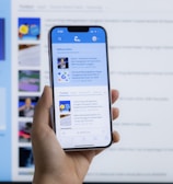 A hand holding a smartphone displaying a news website, with a blurred large computer screen in the background also showing similar content. The webpage on the phone features articles with images and text.