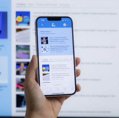 A hand holding a smartphone displaying a news website, with a blurred large computer screen in the background also showing similar content. The webpage on the phone features articles with images and text.