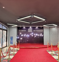 A red carpet leads to a backdrop with bright lights and the words in multiple languages, suggesting a cinematic or award-themed environment. Velvet ropes with gold stanchions line the carpet, and the adjacent wall features movie posters.