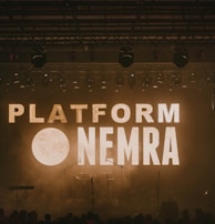 A dimly lit stage with the words 'PLATFORM NEMRA' displayed in large, bold letters across the background screen. Musical instruments such as a keyboard, drums, and microphones are visible on the stage. Stage lights hang from above, contributing to the atmospheric lighting.