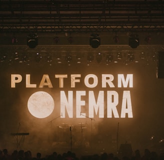 A dimly lit stage with the words 'PLATFORM NEMRA' displayed in large, bold letters across the background screen. Musical instruments such as a keyboard, drums, and microphones are visible on the stage. Stage lights hang from above, contributing to the atmospheric lighting.