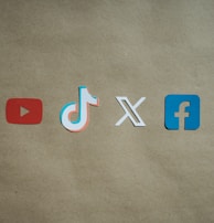 Four social media platform logos are arranged on a brown background. From left to right, the logos are for YouTube in red, TikTok in a combination of red, blue, and white, X in white, and Facebook in blue.